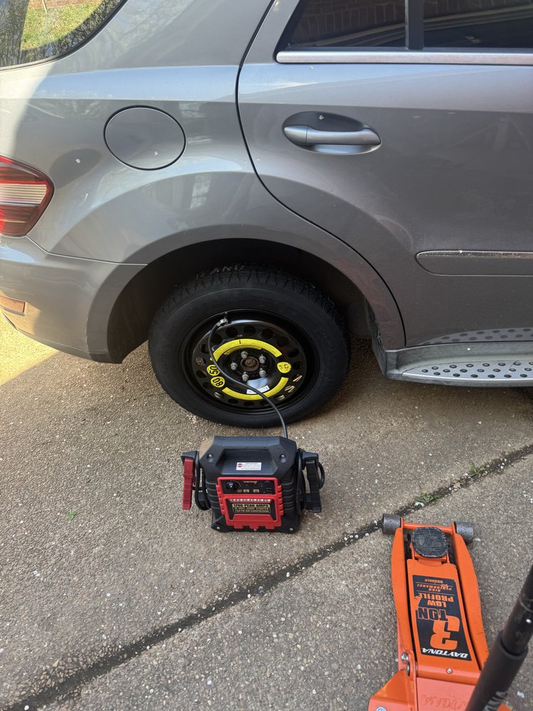 Compact spare tire on a vehicle being inflated with a portable air compressor, floor jack on the driveway